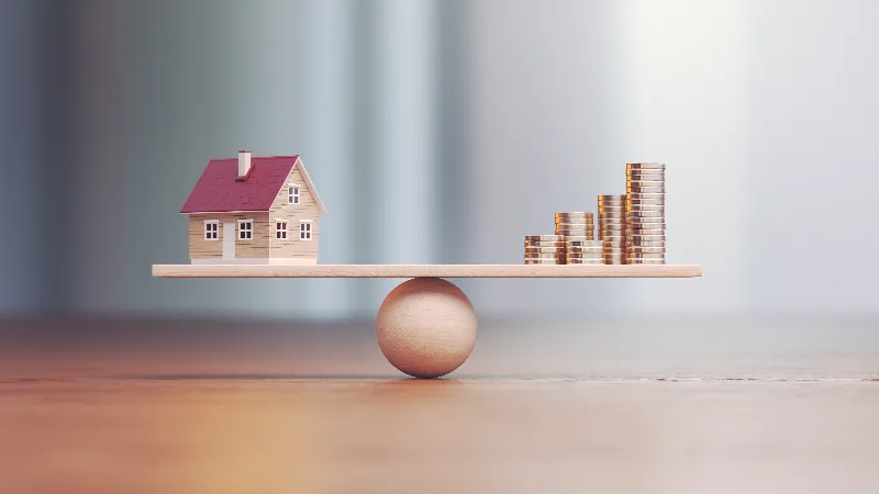 House model and stacked coins balanced on a wooden beam, symbolizing valuation and protection of complex assets in Colorado divorce.